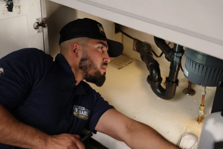 A man in a uniform and cap inspects plumbing under a sink, reaching towards pipes and a garbage disposal unit during a plumbing repair.