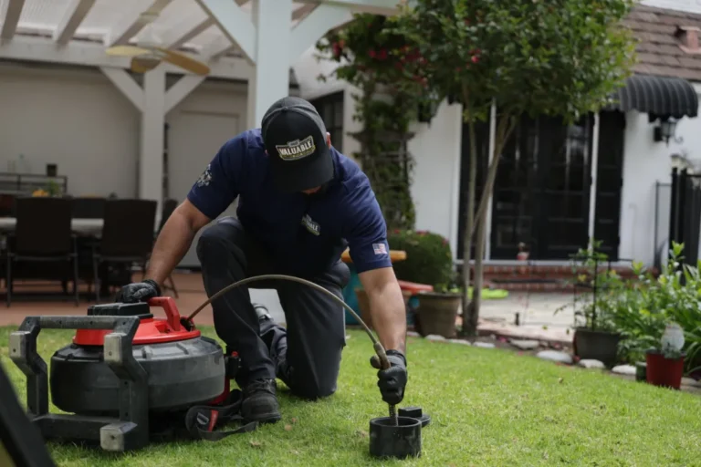 A worker kneels on a lawn, using equipment to treat or inspect the grass in a residential backyard, while also checking for any drain cleaning needs.