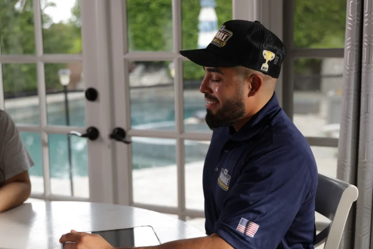 A man in a navy uniform and hat sits at a table with a tablet, near large windows overlooking a pool while discussing kitchen plumbing installation.