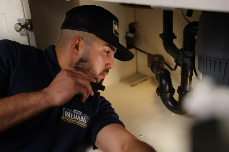 A man wearing a uniform and cap conducts a plumbing inspection, carefully checking under a sink with a flashlight.