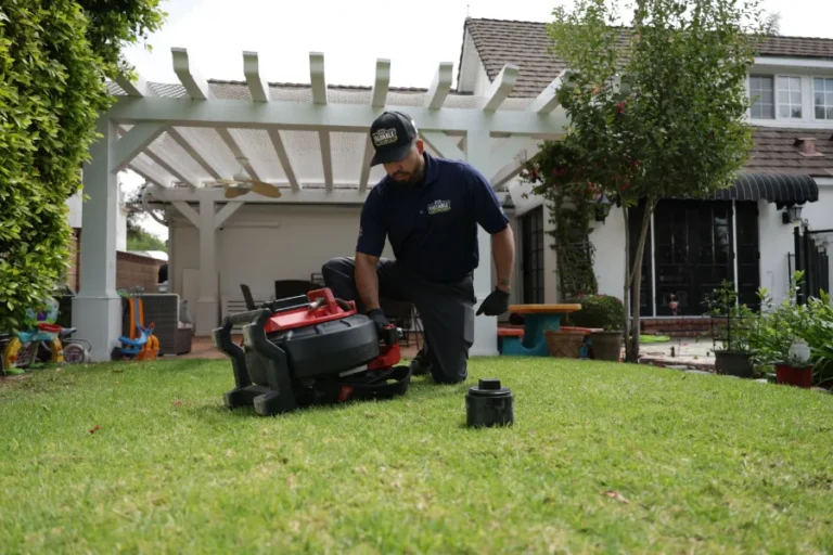 A man kneels on a lawn, inspecting a robotic lawn mower in a backyard with a pergola and house in the background—considering if advanced tools like hydro jetting service could simplify other home maintenance tasks.