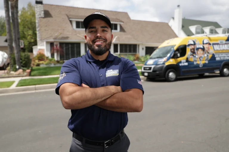 A man in a navy blue polo shirt and cap stands with arms crossed on a suburban street, ready to provide expert tankless water heater repair, with a branded service van and houses in the background.