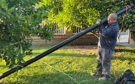 A man in work gloves holds and positions a long black pipe for burst pipe repair in a grassy yard, surrounded by trees and gardening tools.