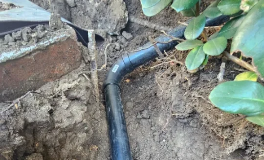 A black plastic pipe, part of a sewer line installation, runs underground next to a brick wall and plants, with soil and roots visible around the exposed section.