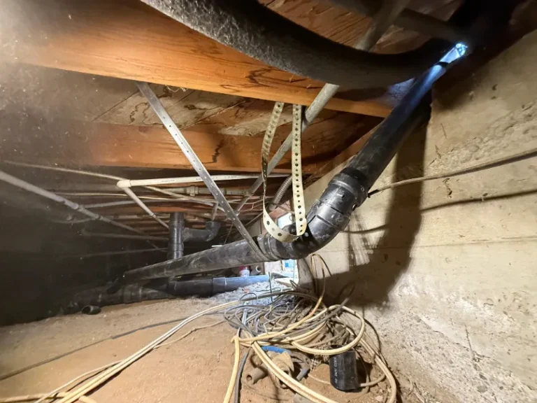 Crawl space beneath a building featuring exposed pipes, plumbing installation, wires, wooden beams, and a concrete wall. Natural light enters from a small opening.