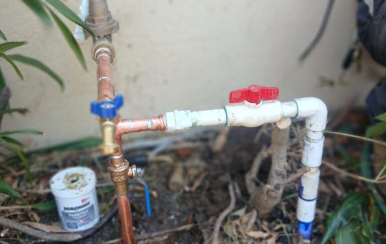 Outdoor water line repair setup featuring copper and PVC pipes, two shut-off valves, and a can of PVC primer or adhesive, positioned by a beige wall and surrounded by plants.