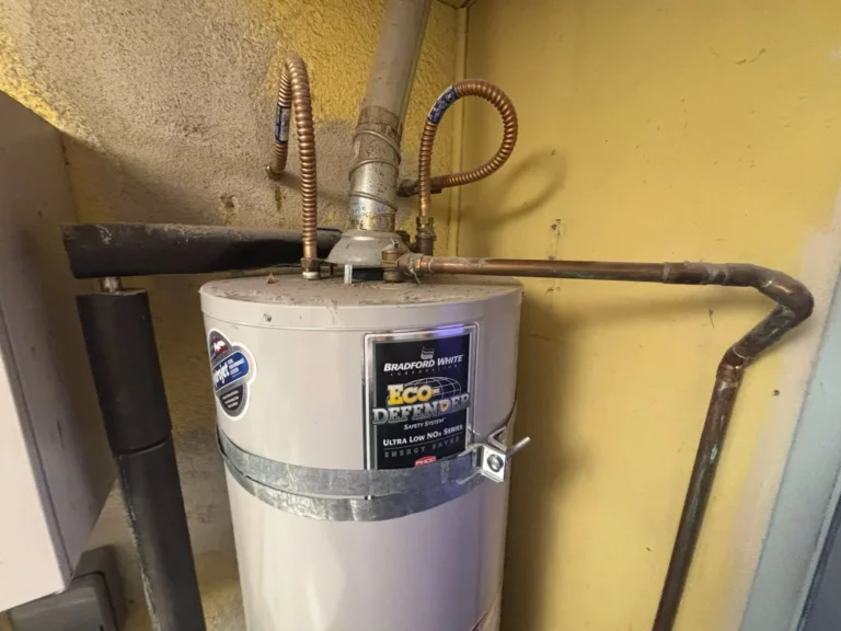 A Bradford White Eco Defender water heater is installed against a yellow wall, with various pipes connected at the top and sides, highlighting the importance of regular water heater maintenance.