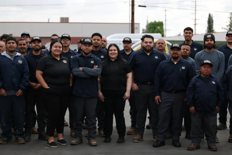 A group of workers, both men and women, stand together outdoors; most are wearing matching navy uniforms and caps, while two women stand in the center in black shirts.