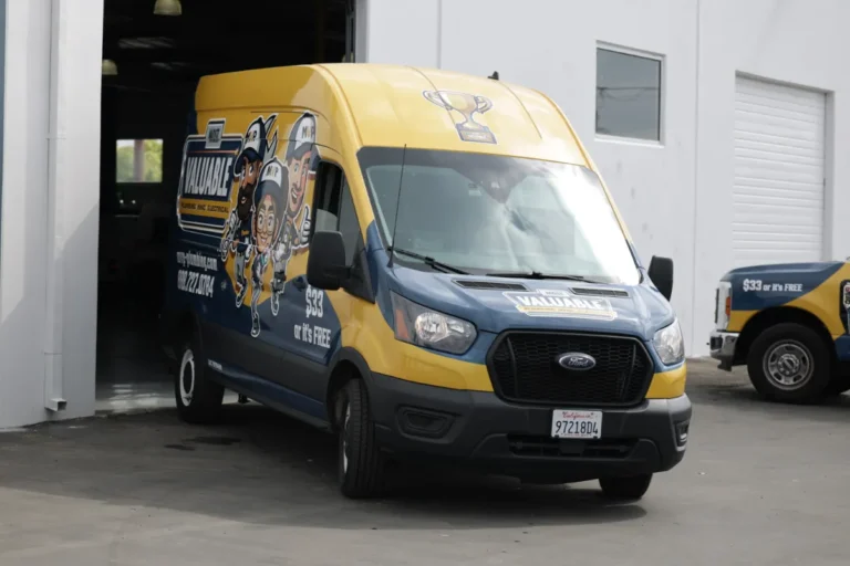 A blue and yellow delivery van with cartoon graphics and a trophy illustration is parked outside a garage next to another similarly designed vehicle.