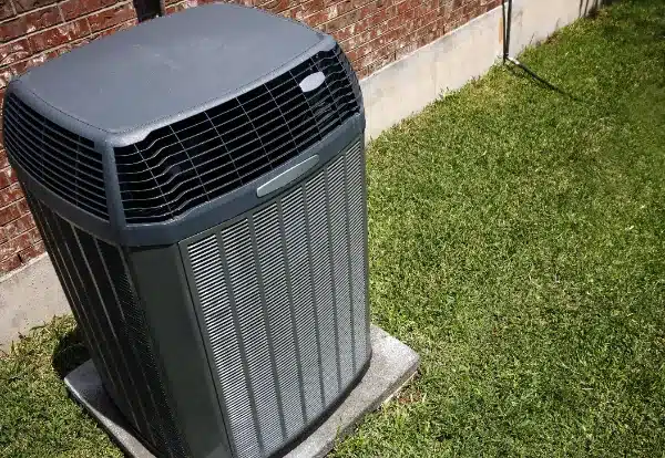 An outdoor air conditioning unit is placed on a concrete slab beside a brick wall, surrounded by a grassy lawn.