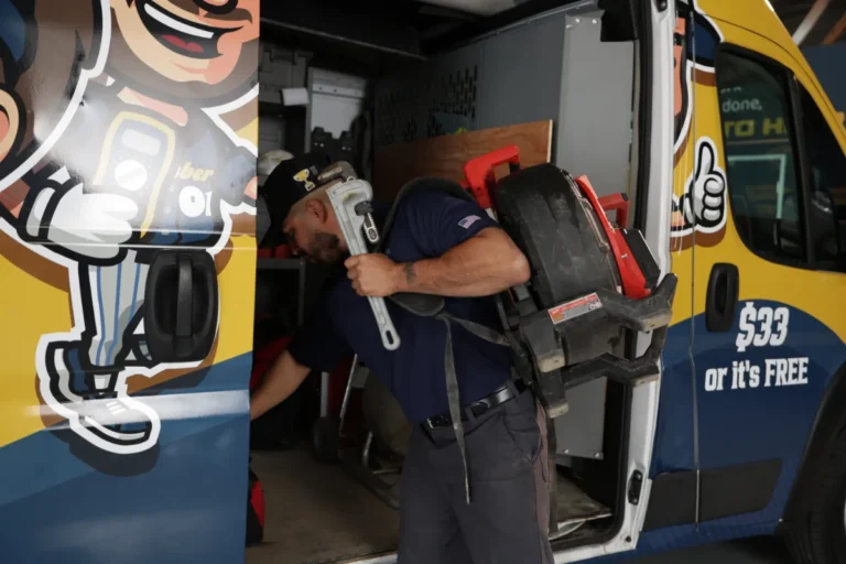 A plumber with tools enters a branded service van, carrying a pipe wrench and drain cleaning machine.