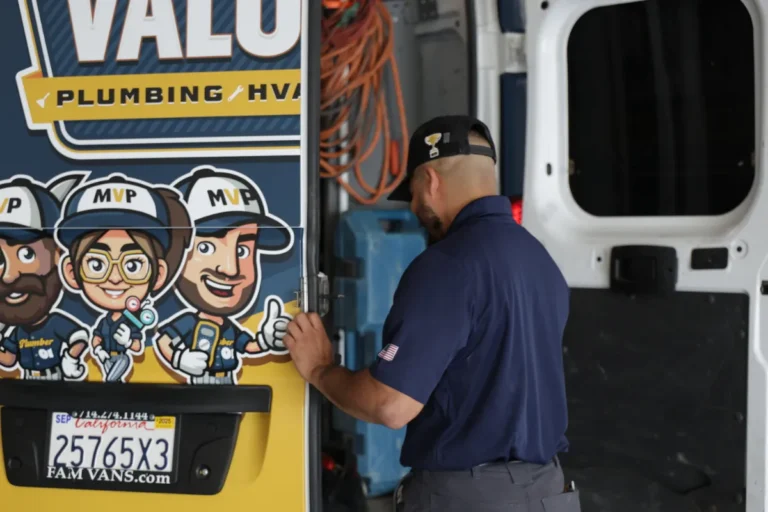 A man in a navy uniform stands at the back of a plumbing service van with cartoon graphics and an open door, preparing equipment.