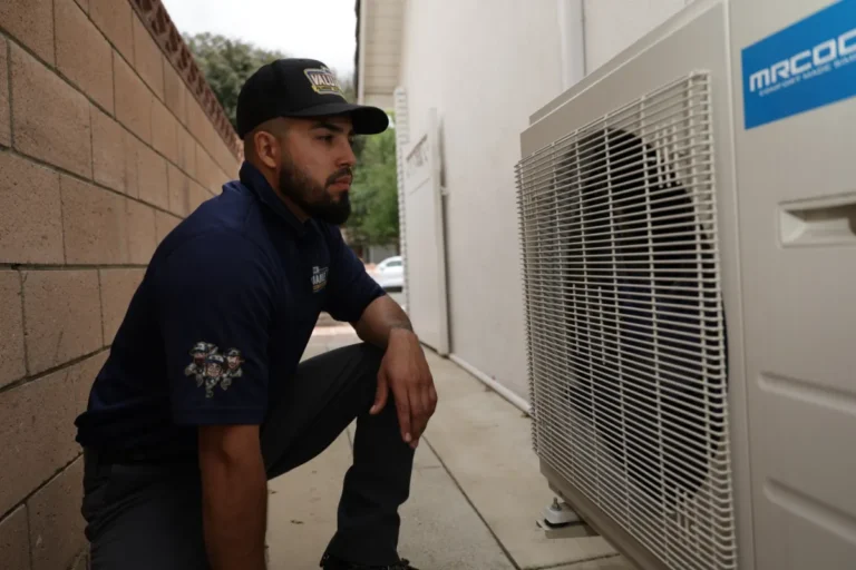 A man wearing a uniform and hat kneels next to an outdoor air conditioning unit beside a building.