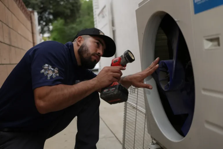A man wearing a navy shirt uses a power drill to repair or inspect the inside of a washing machine outdoors.