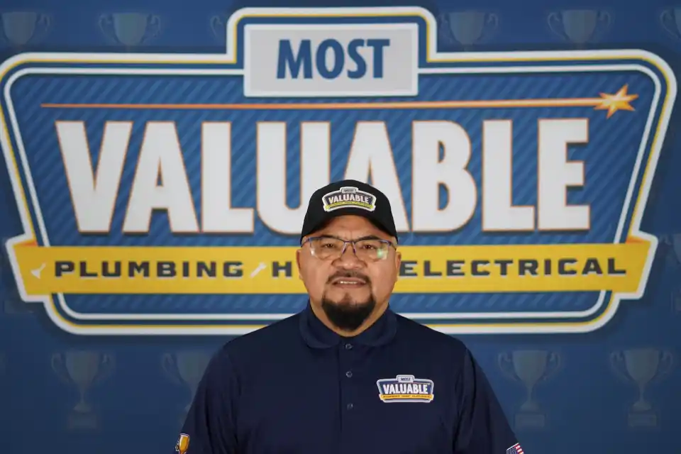 Meet the team: A man wearing glasses and a “Valuable” hat and shirt stands in front of a sign reading "Most Valuable Plumbing HVAC Electrical.