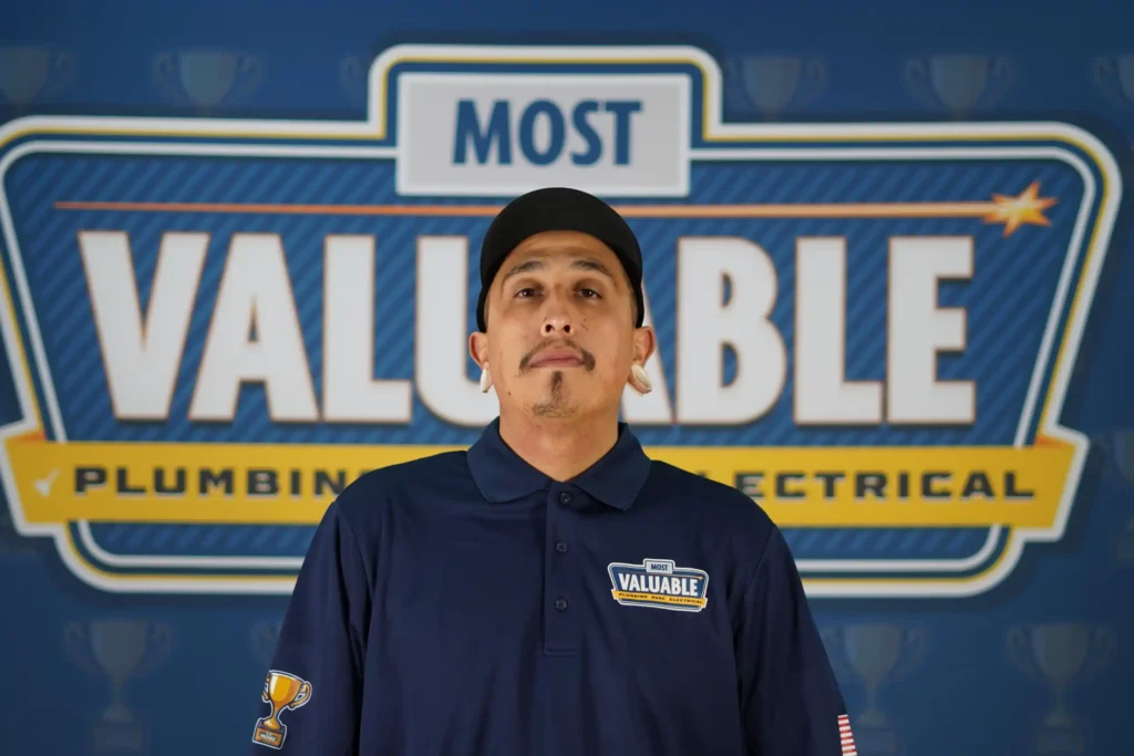 A man wearing a "Most Valuable" uniform stands in front of a sign that reads "Most Valuable Plumbing Electrical," ready to welcome you as you meet the team.