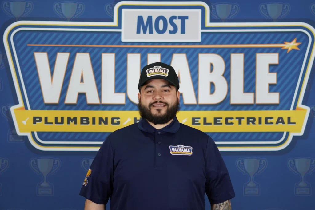 A man in a "Most Valuable" uniform and hat stands in front of a sign that reads "Most Valuable Plumbing and Electrical," ready to welcome you as you meet the team.