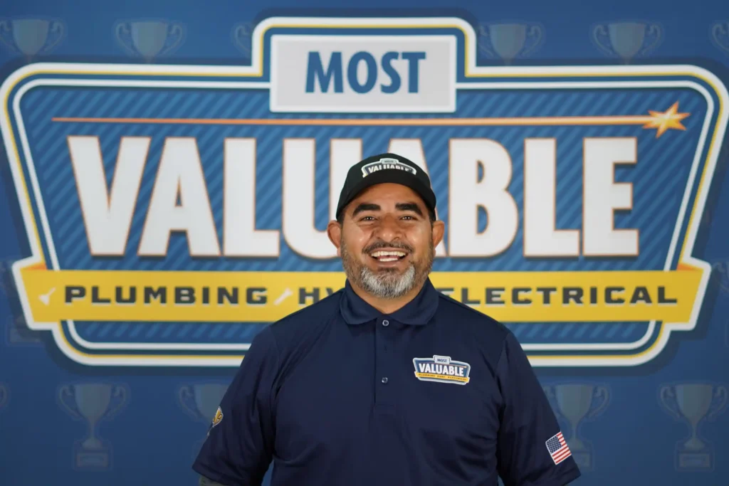 Meet the team: A man in a navy uniform and cap stands smiling in front of a "Most Valuable Plumbing HVAC Electrical" sign with trophy symbols in the background.