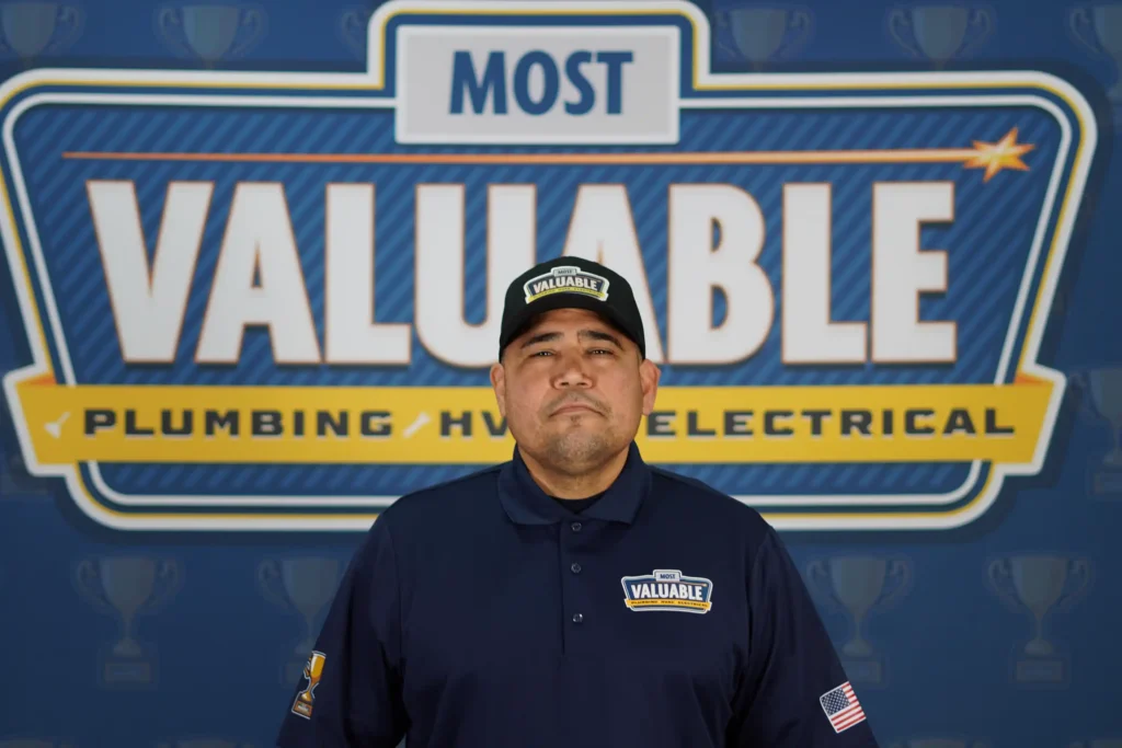 A man in a "Most Valuable" uniform stands in front of a sign that reads "Most Valuable Plumbing HVAC Electrical." Meet the team dedicated to providing outstanding service and expertise for all your home needs.