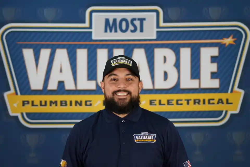 A man wearing a "Most Valuable" hat and shirt stands smiling in front of a sign that reads "Most Valuable Plumbing & Electrical"—ready to welcome you and help you meet the team.