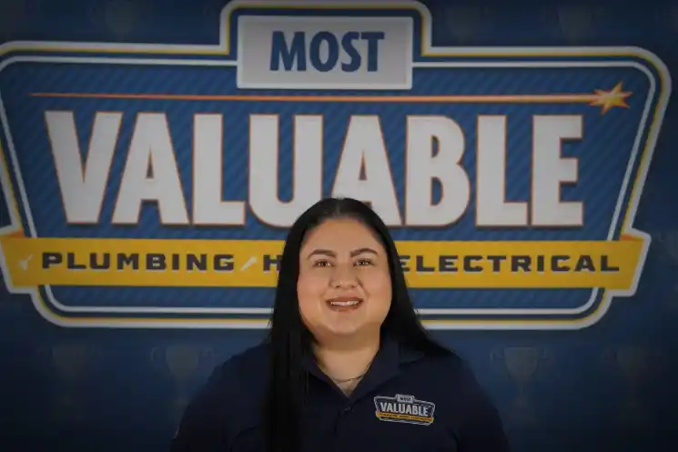A woman in a navy shirt stands smiling in front of a sign that reads "Most Valuable Plumbing Heating Electrical"—proudly inviting you to meet the team behind the service.
