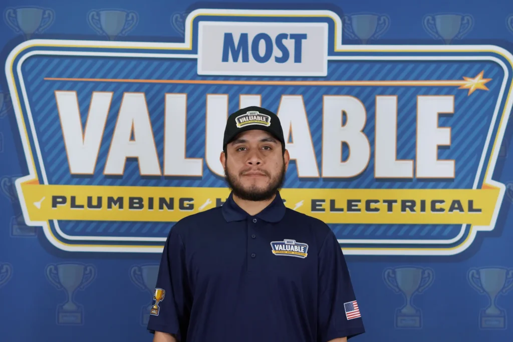A man in a navy uniform and cap stands in front of a backdrop that reads "Most Valuable Plumbing Electrical," ready to meet the team behind exceptional service.