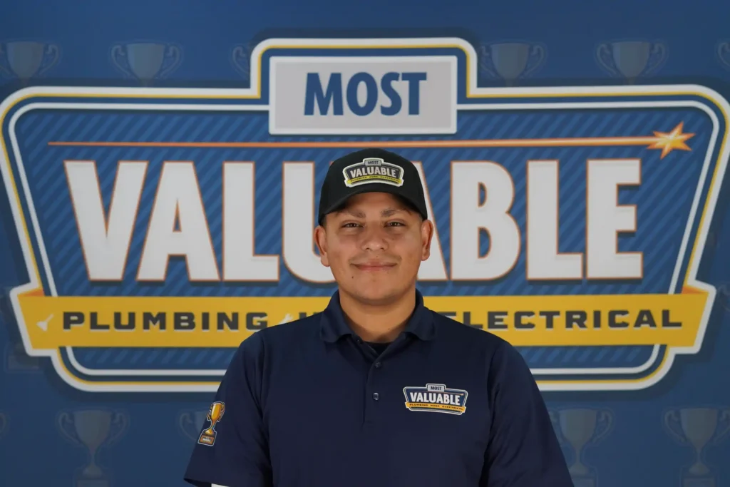 A person wearing a "Most Valuable" uniform and hat stands in front of a sign that reads "Most Valuable Plumbing and Electrical." Meet the team behind your trusted local service experts.