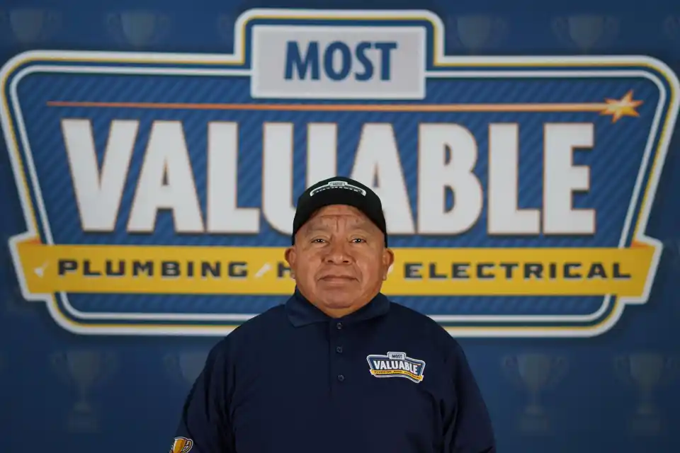 A man wearing a "Most Valuable" uniform stands in front of a sign that reads "Most Valuable Plumbing and Electrical," ready to greet you as part of the meet the team experience.