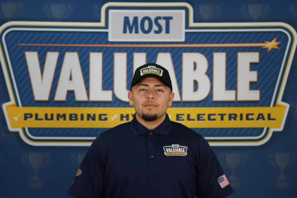 A man in a "Most Valuable" uniform and hat stands in front of a sign that reads "Most Valuable Plumbing HVAC Electrical," ready to welcome you to meet the team.