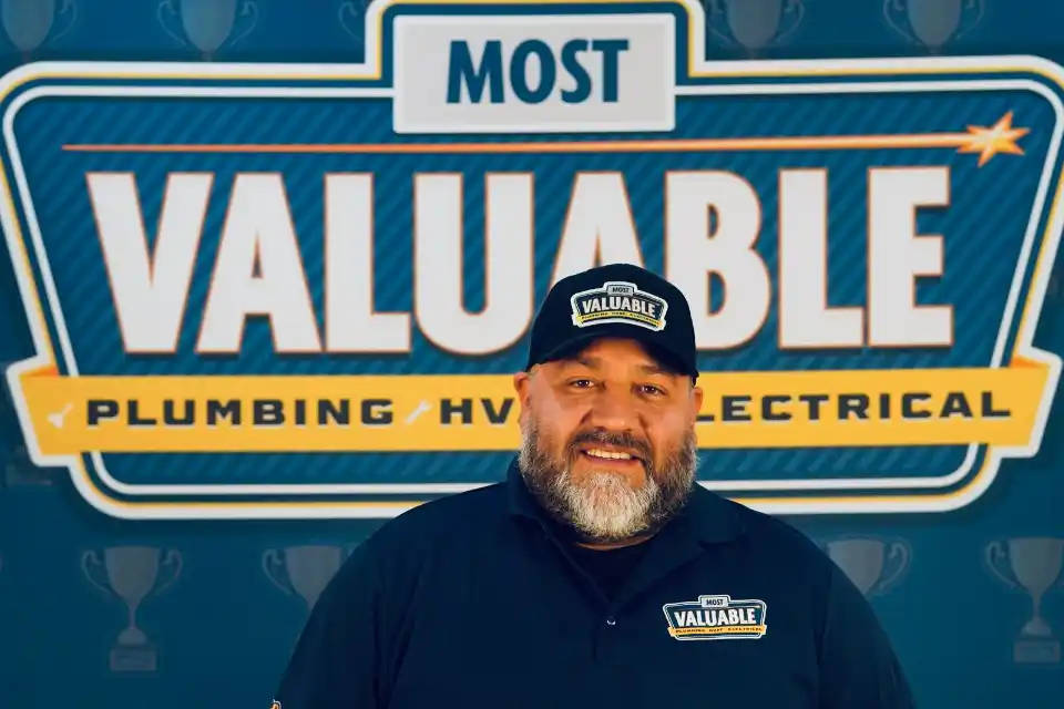 A man wearing a "Most Valuable" hat and shirt stands in front of a sign that reads "Most Valuable Plumbing / HVAC / Electrical," ready to welcome you as part of the meet the team experience.