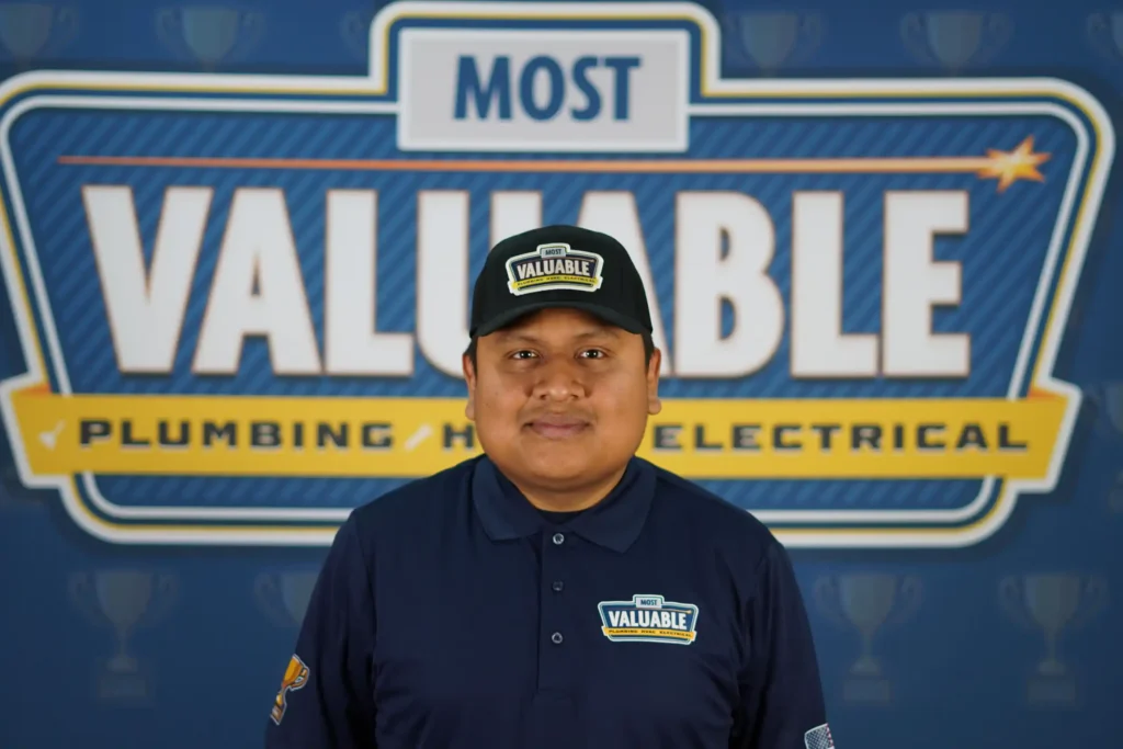 A man in a navy "Most Valuable" uniform and hat stands in front of a backdrop with the words "Most Valuable Plumbing Heating Electrical." Meet the team behind your trusted home services.