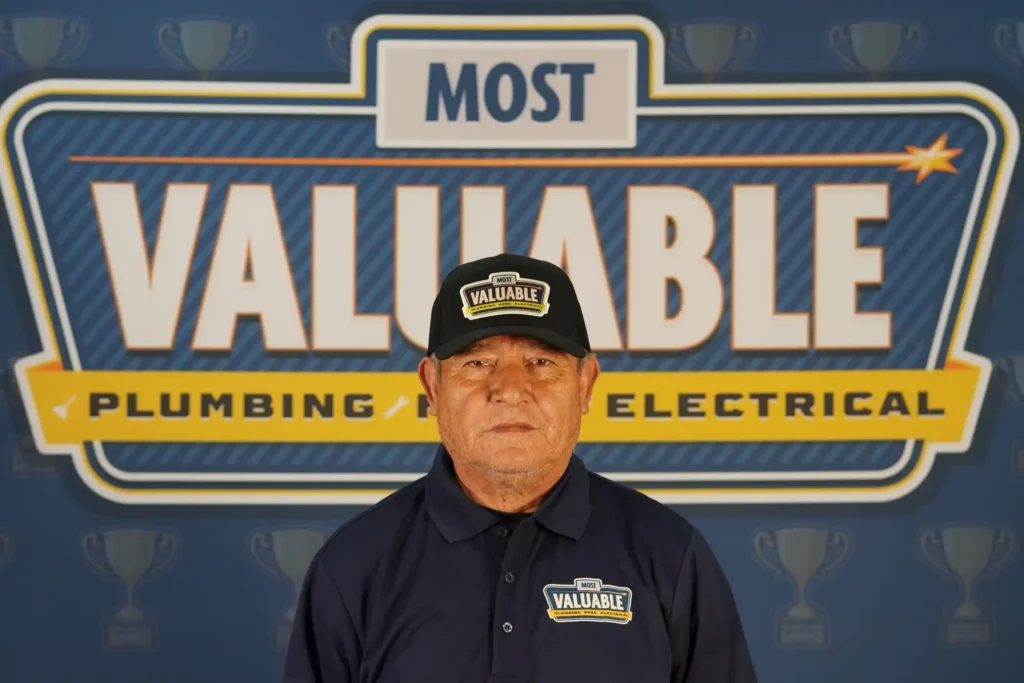 A man wearing a "Most Valuable" hat and shirt stands in front of a sign that reads "Most Valuable Plumbing Electrical," with trophy icons in the background—meet the team behind your trusted service experts.