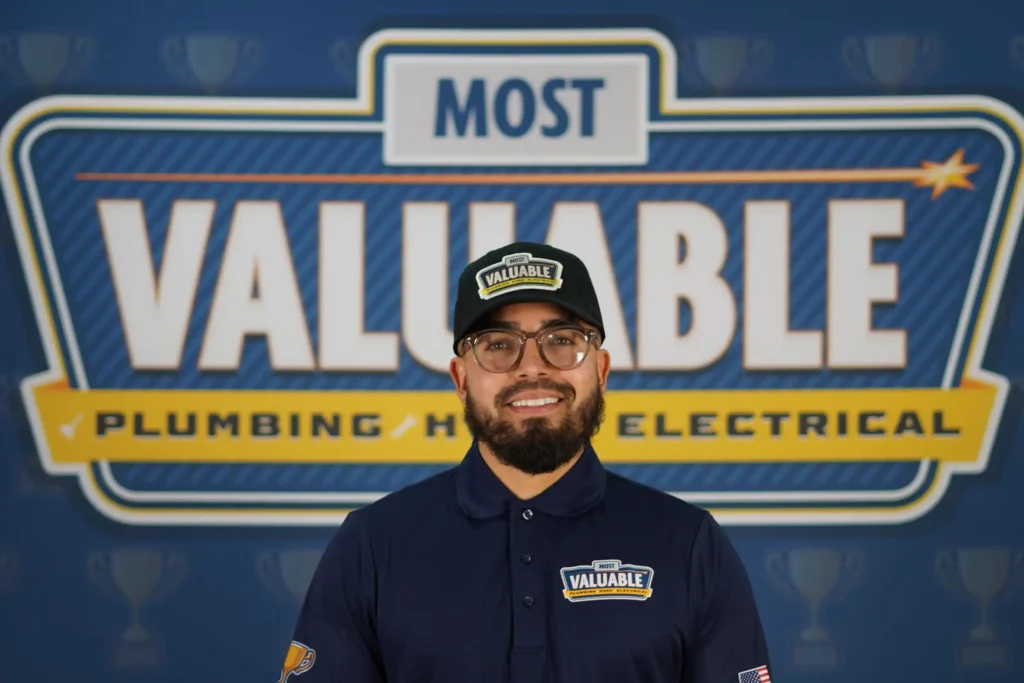 A man wearing a "Most Valuable" hat and shirt stands smiling in front of a large "Most Valuable Plumbing HVAC Electrical" sign—proudly representing his role as part of the meet the team crew.