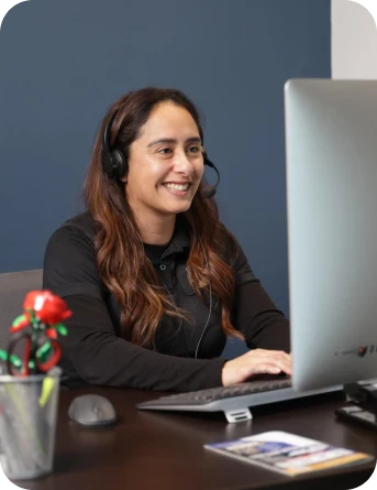 A woman wearing a headset sits at a desk, smiling while working on a computer. A pen holder with a flower and some papers are on the desk.