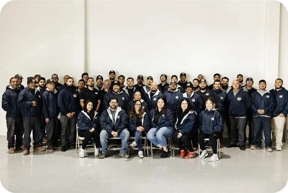 A large group of people pose for a group photo indoors against a plain white wall. Some are seated in front while others stand behind them.