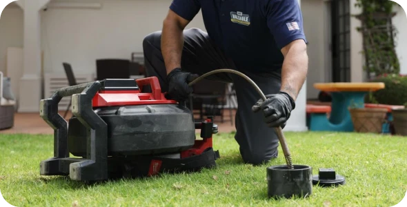A person wearing gloves kneels on grass, using a red and black drain cleaning machine with a flexible hose attachment.