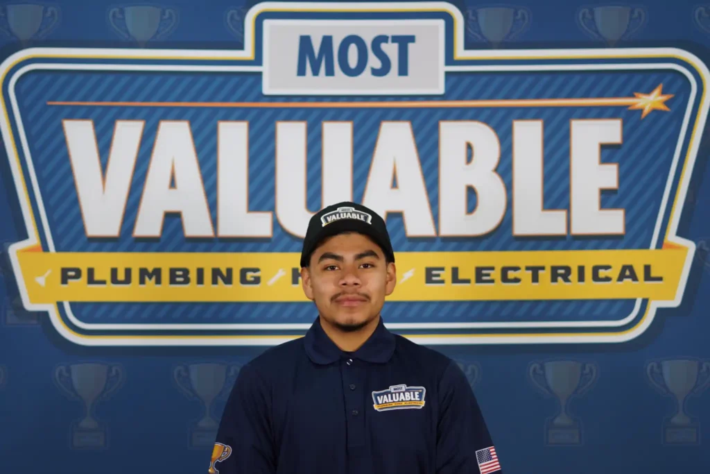 A man in a blue uniform and cap stands in front of a sign that reads "Most Valuable Plumbing Electrical," ready to meet the team behind your trusted service.