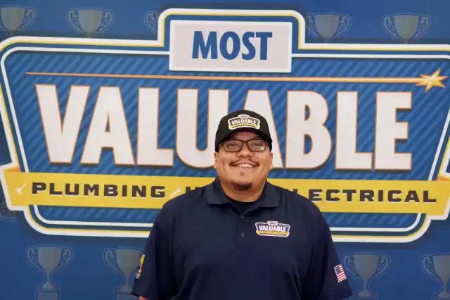 Meet the team: A man in a navy uniform and cap stands smiling in front of a sign that reads "Most Valuable Plumbing and Electrical.