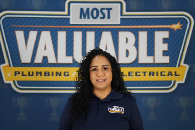 A woman with curly hair stands in front of a sign that reads "Most Valuable Plumbing & Electrical," wearing a matching branded shirt—ready to help you meet the team.