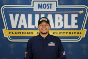 A man wearing a "Most Valuable" hat and shirt stands in front of a sign that reads "Most Valuable Plumbing Heating Electrical," ready to help you meet the team behind your trusted home services.