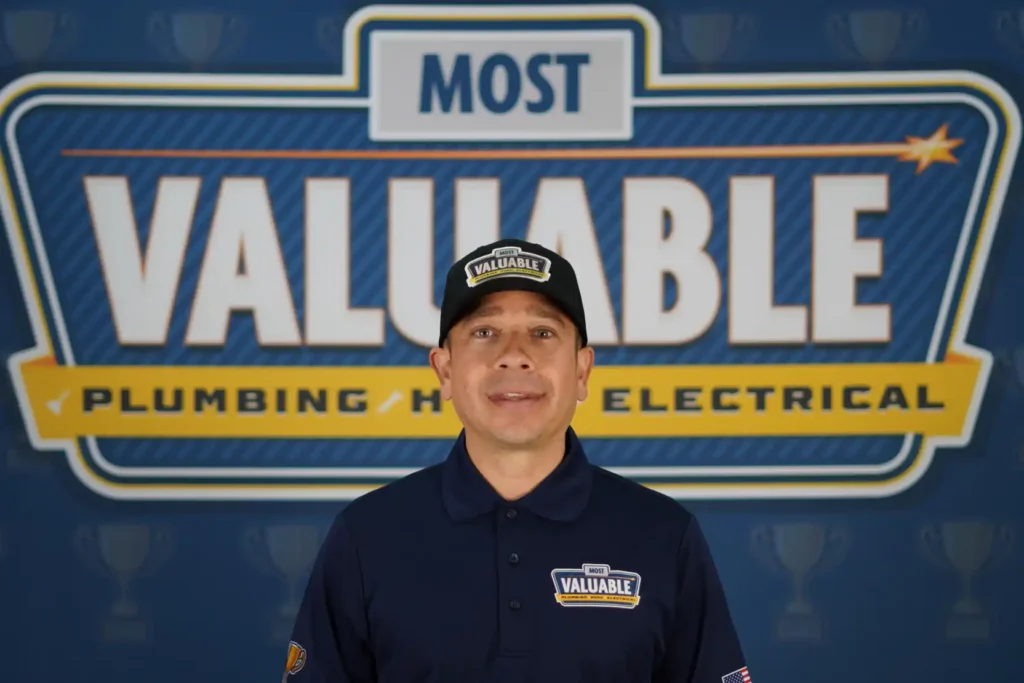 Man wearing a "Most Valuable" uniform and hat stands in front of a large sign that reads "Most Valuable Plumbing HVAC Electrical," ready to introduce you to the team. Come meet the team behind your trusted home services.