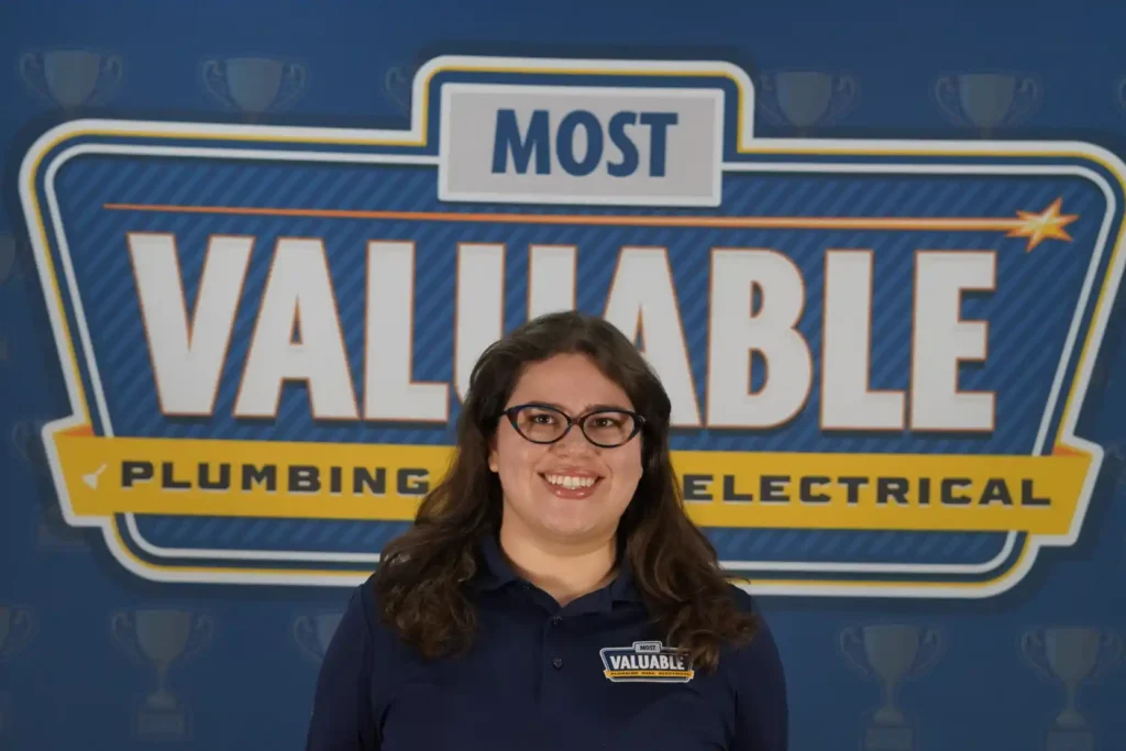 A woman in a "Most Valuable" polo shirt stands smiling in front of a sign reading "Most Valuable Plumbing Electrical," ready to help you meet the team.
