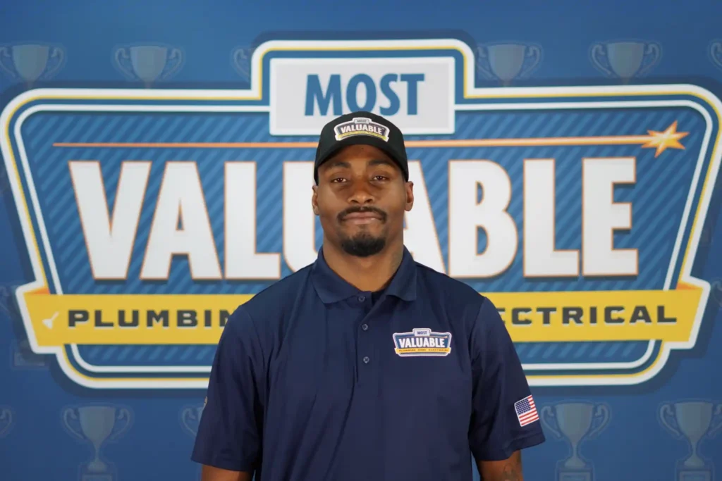 Meet the team: A man in a navy blue uniform and cap stands in front of a "Most Valuable Plumbing Electrical" sign with trophy graphics in the background.