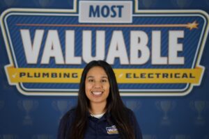 A woman in a navy shirt stands smiling in front of a large sign reading "Most Valuable Plumbing Electrical," ready to introduce you to the Meet the Team experience.