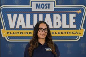 Meet the team: A woman wearing glasses and a “Most Valuable” polo shirt stands in front of a sign that says “Most Valuable Plumbing Electrical.”.