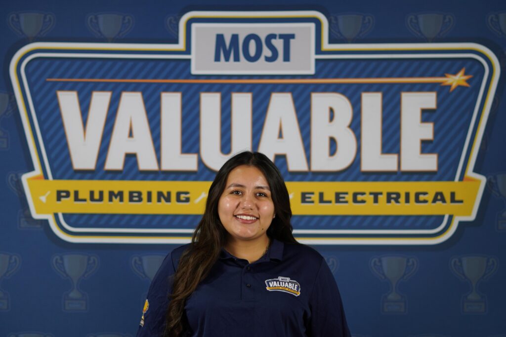 A woman in a navy "Valuable" shirt stands smiling in front of a sign that reads "Most Valuable Plumbing Electrical"—a great way to meet the team behind the company.