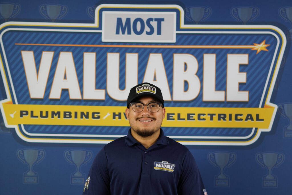 Meet the team: A person wearing glasses and a "Most Valuable" hat and shirt stands smiling in front of a "Most Valuable Plumbing HVAC Electrical" sign.
