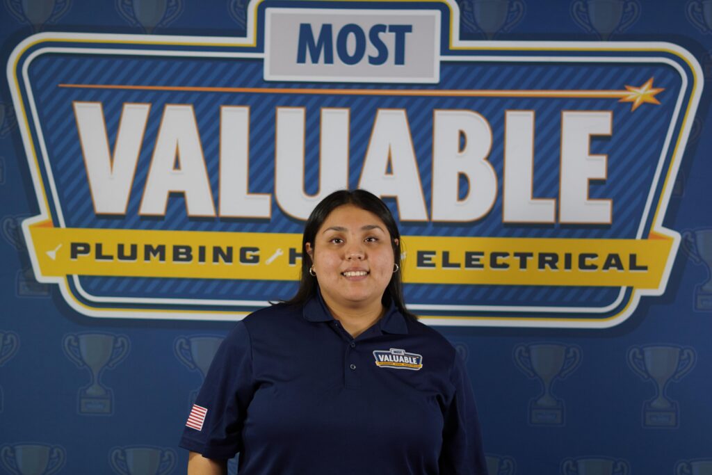 A person in a navy blue "Most Valuable" logo shirt stands in front of a "Most Valuable Plumbing Heating Electrical" backdrop, ready to meet the team and showcase their expertise.