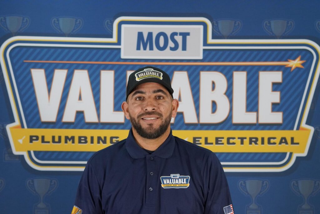 A man in a navy uniform and hat stands in front of a sign reading "Most Valuable Plumbing & Electrical," ready to welcome you as part of our meet the team spotlight.
