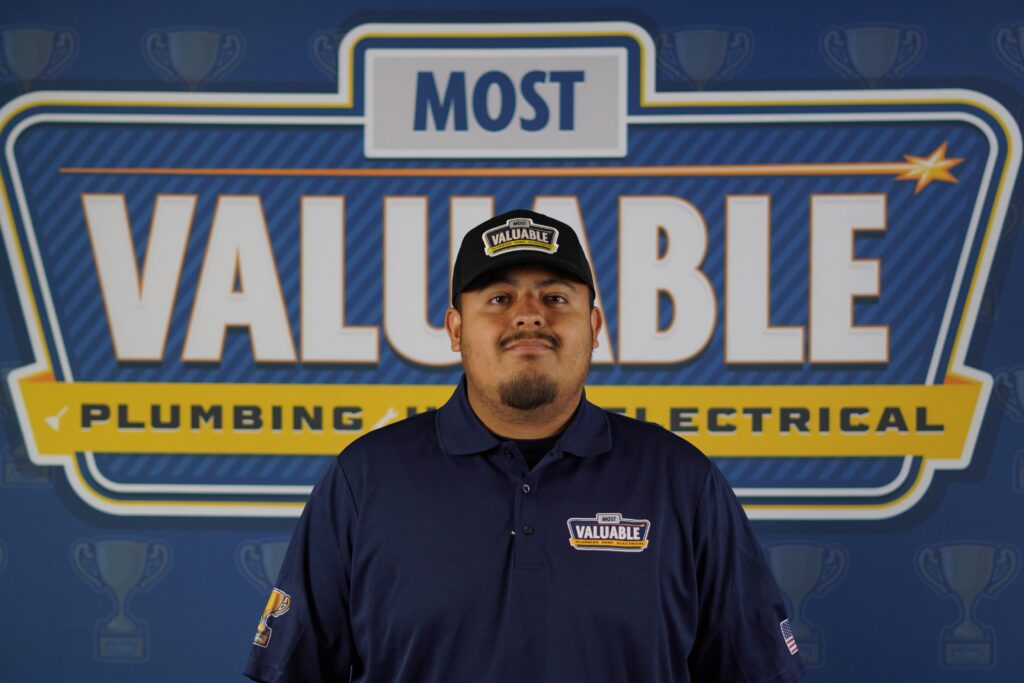 Meet the team: A man in a dark blue "Most Valuable" uniform and cap stands in front of a large sign that reads "Most Valuable Plumbing and Electrical.